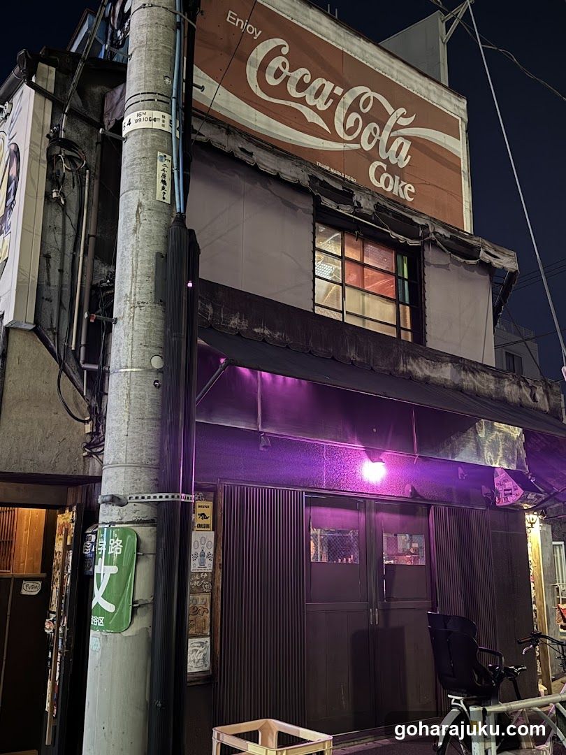 A nighttime photo of a shop front with a Coca-Cola sign above the entrance. The building has a purple awning and a wooden door. A bicycle is parked nearby, and there are various signs and posters on the wall.