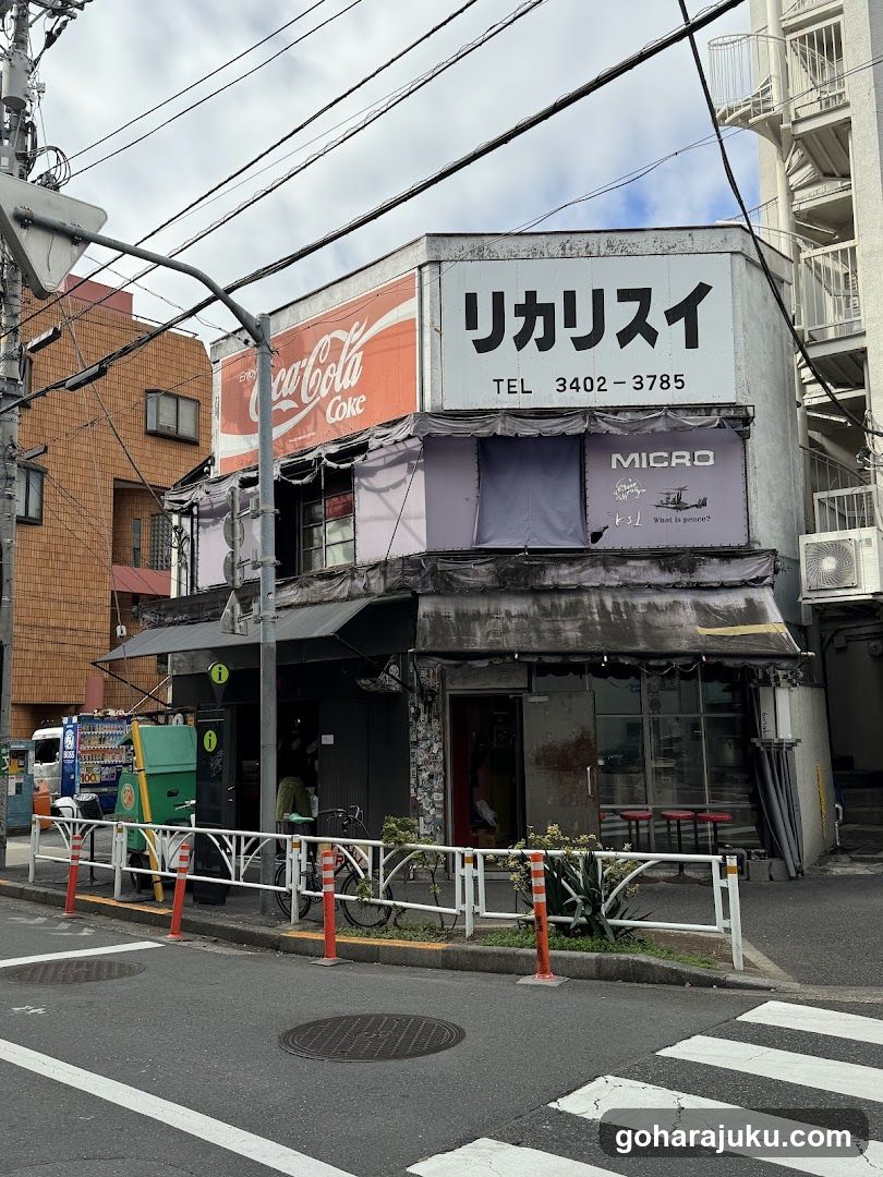 A corner shop with a Coca-Cola sign and Japanese signage reading 'リカリスイ' (Rikarisui). The storefront features a dark awning, glass windows, and outdoor seating. A bicycle is parked nearby, and the building has visible wear.