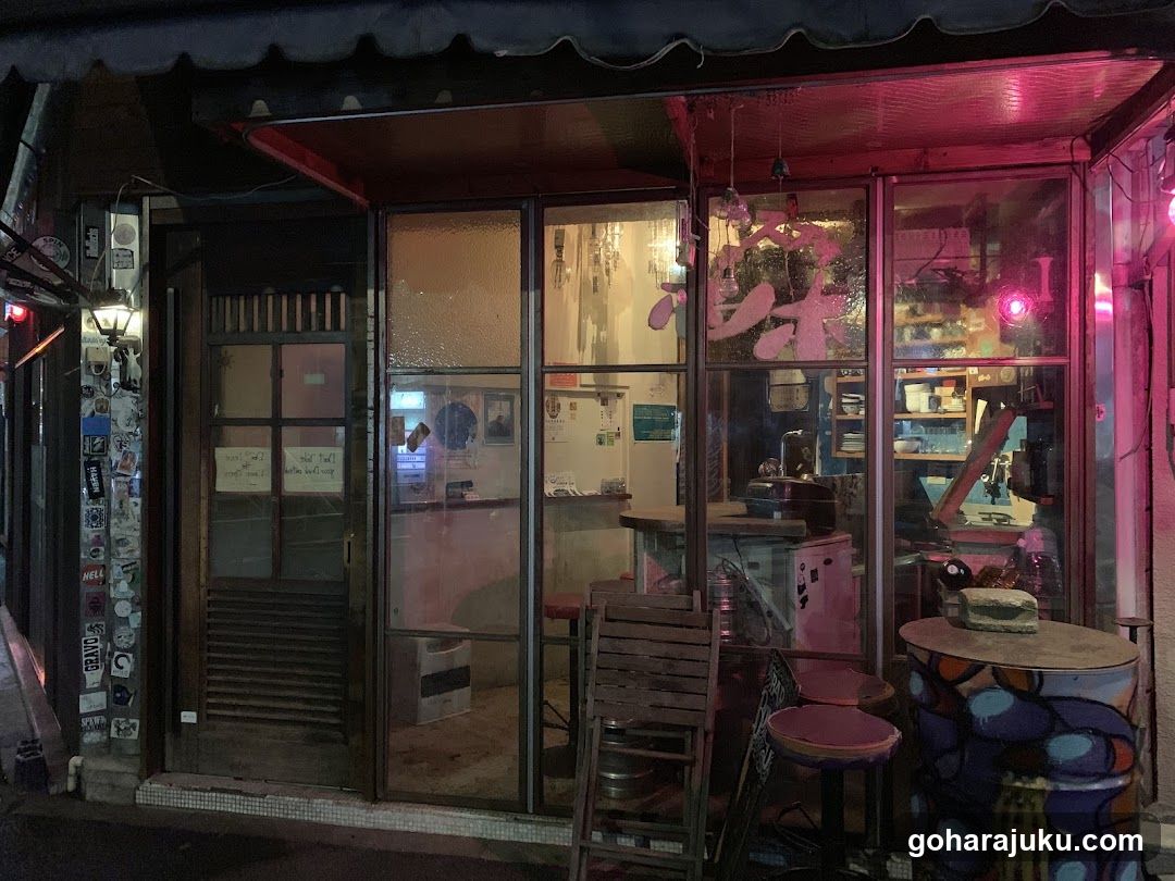 A shop entrance at night featuring glass doors and windows, illuminated by pink neon lights. Visible items include a wooden chair, colorful barrel table, and interior shelves with various goods. Stickers adorn the wall to the left.