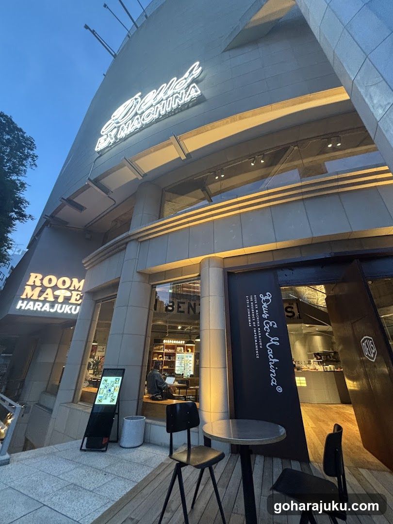 Evening shot of the exterior facade showing illuminated signage reading 'Diner & MACHINA' and 'ROOM MATE HARAJUKU'. A black entrance door displays vertical white script text. Outside seating includes a small round table with two chairs on wooden decking. A digital menu board stands near the sidewalk, displaying colorful graphics.