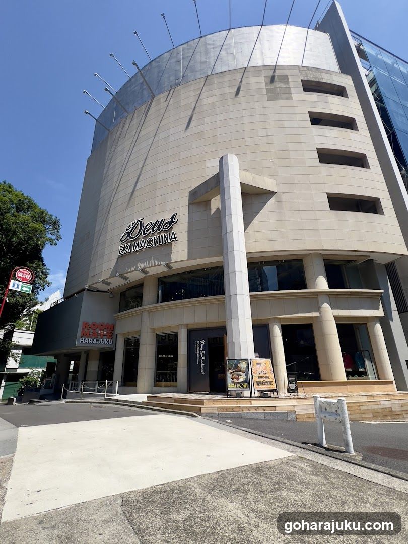 A modern, curved building facade with 'Deus Ex Machina' signage above the entrance and 'HARAJUKU' below. Large glass windows display interior lighting. A vertical column supports an overhang. Street signs and a white barrier are visible in front.
