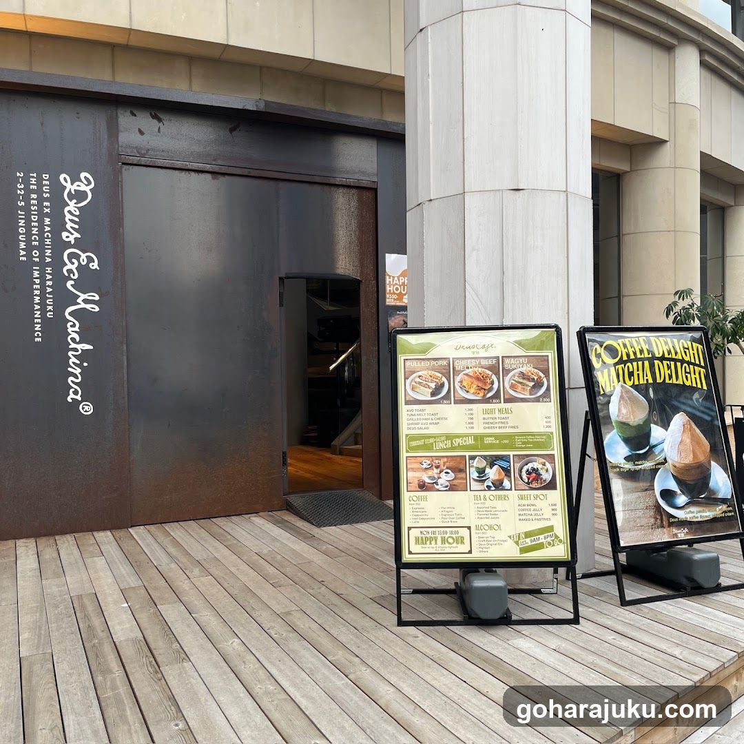Exterior of a modern coffee shop with dark metal entrance and wooden deck. Two menu boards stand outside, one listing food items like 'Chicken Burger' and 'Matcha Latte', the other featuring matcha desserts.