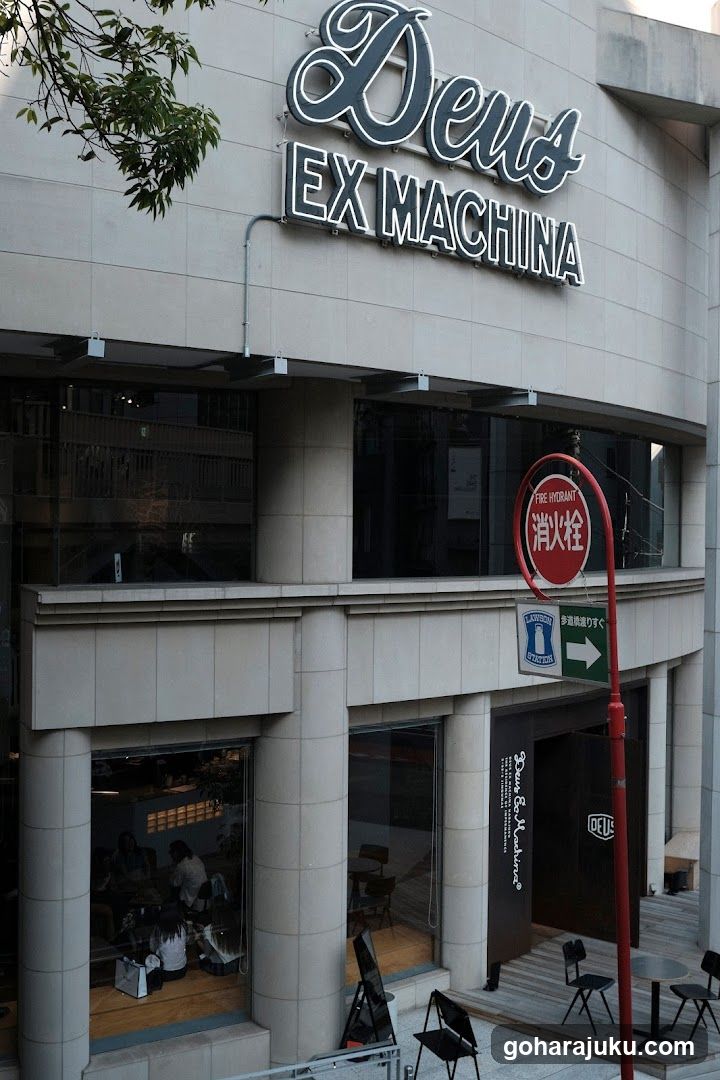 The image shows the exterior of a building with 'Deus EX MACHINA' signage prominently displayed on the facade. The entrance features large glass windows and doors, allowing visibility into the interior where people are seated at tables. A red fire extinguisher sign is mounted on a pole near the entrance.