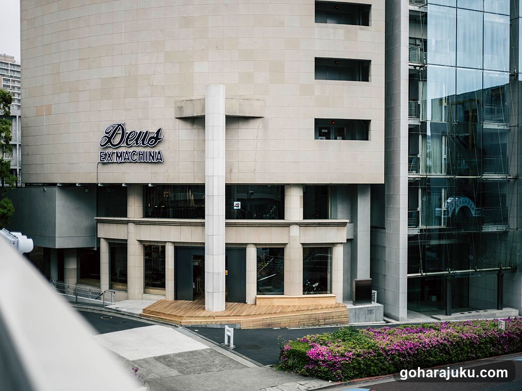 Photo of the physical exterior entrance and storefront facade of a building with visible signage reading 'Deus EX MACHINA' on the upper wall. The ground floor features large glass windows, columns, and a wooden platform leading to the entrance. Adjacent is a modern glass structure. No menus or printed materials are visible.