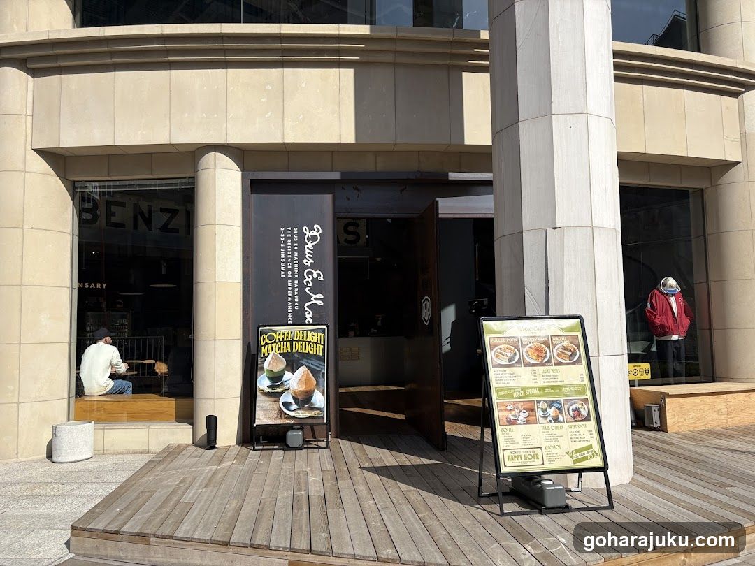 Exterior view of a coffee shop with beige stone facade and large glass windows. A vertical sign reads 'BENZU' on the left window, while another displays 'COFFEE & CAFE'. Two freestanding menu boards are placed outside: one advertises 'COFFEE & CAKE', showing images of pastries; the other lists food items like rice bowls and sandwiches with prices in yen.