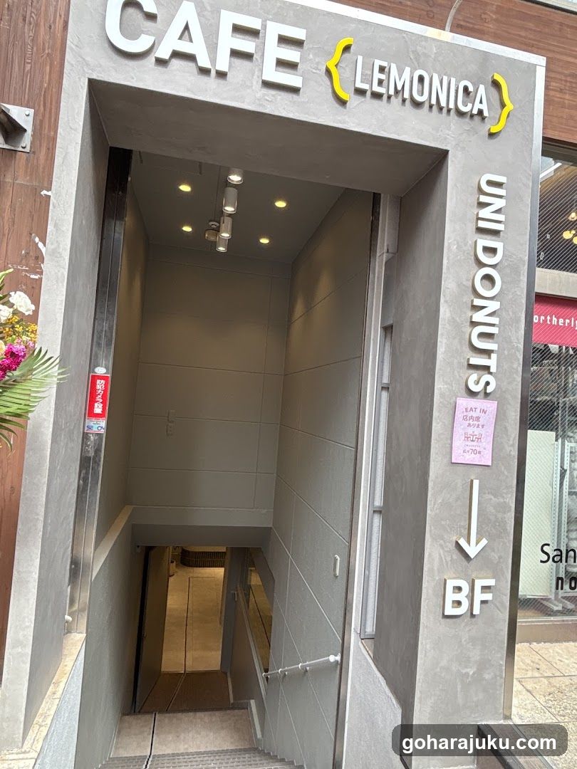 Exterior view of Cafe Lemonica entrance, featuring a modern concrete facade with signage reading 'CAFE LEMONICA' and 'UNIDONUTS BF'. A pink notice is posted on the wall, indicating restricted entry or hours.