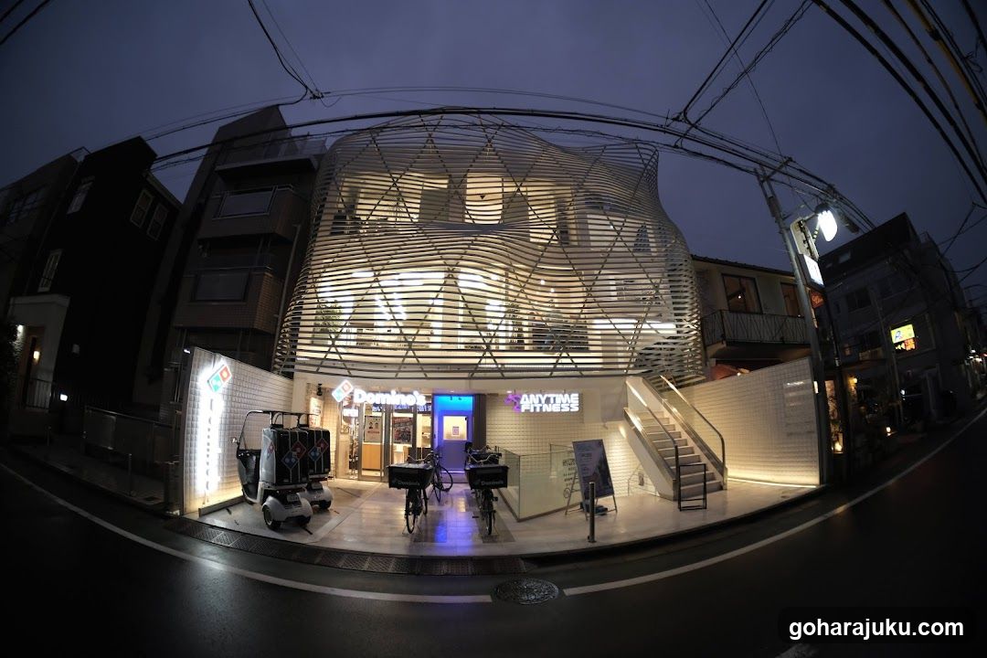 Fisheye view of a modern fitness center entrance with illuminated geometric facade and signage reading 'AIKUTSU FITNESS'. Bicycles parked outside, adjacent to a convenience store entrance.