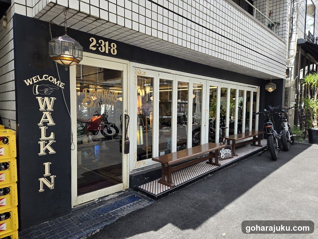 The image shows the exterior of a coffee shop with a black and white tiled facade. The address '2-318' is displayed above the entrance. A sign on the left reads 'WELCOME WALK IN'. There are wooden benches outside, and several motorcycles parked nearby. Inside, through the glass doors, a red motorcycle is visible.