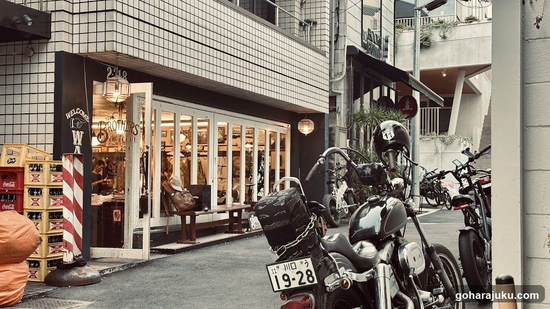 A shop with a tiled facade and large glass windows, displaying interior items like clothing or accessories. A red-and-white striped pole stands outside near stacked crates of Coca-Cola bottles. Motorcycles are parked in front.