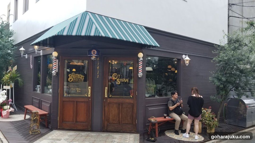 A dark wooden storefront with a green and white striped awning. Two glass doors feature gold lettering reading 'BROTHERS BARBERSHOP' and 'CUT SHAVE'. A barber pole sign is mounted on the wall beside the entrance. Two people sit on benches outside near potted plants.