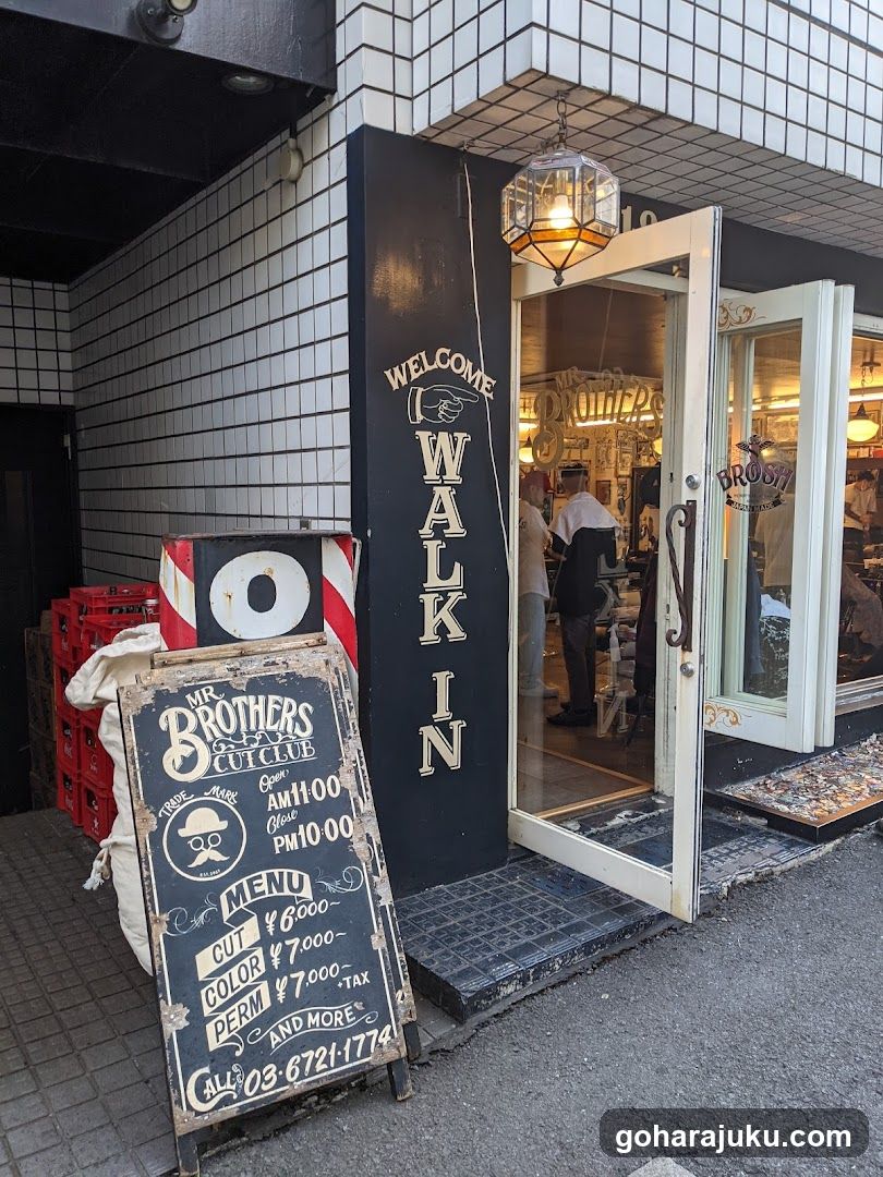 A barbershop entrance with a black vertical sign reading 'WELCOME WALK IN' and a chalkboard menu board displaying services like cut, color, perm, and pricing.