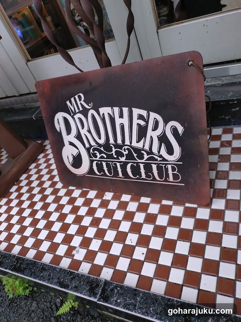 A weathered, rusty metal sign reading 'MR BROTHERS CUT CLUB' in stylized white lettering is mounted on a tiled surface outside a shop entrance. The tiles are arranged in a brown and white checkerboard pattern. Above the sign, twisted metal branches or vines hang down near glass doors.
