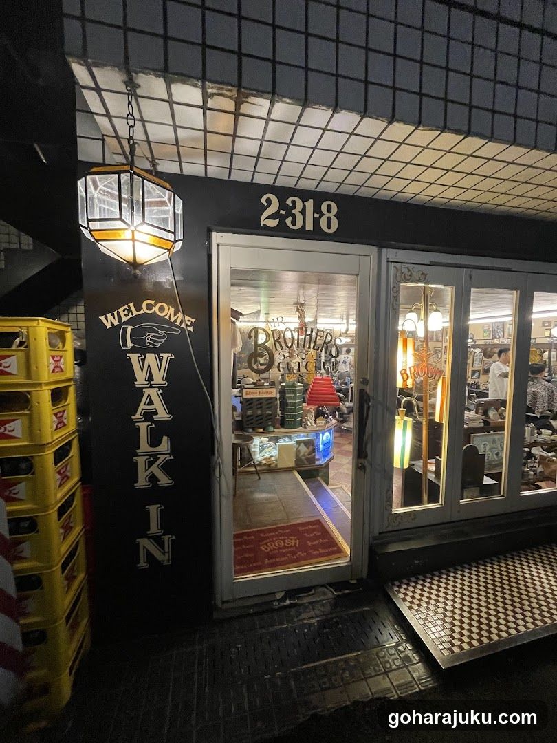 A storefront with a black facade and white grid ceiling above. A hanging lantern illuminates the entrance. Yellow beer crates are stacked to the left. The address '2-31-8' is displayed above the door. Inside, shelves hold various items including lamps and decor. A red welcome mat reads 'WELCOME'. Glass doors show interior lighting and merchandise.
