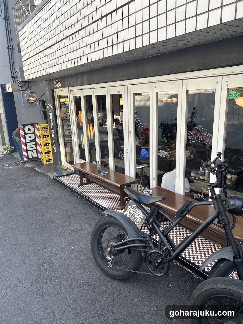 A street-level view of a bar and grill with large glass windows, wooden benches outside, an open sign, and a bicycle parked in front.