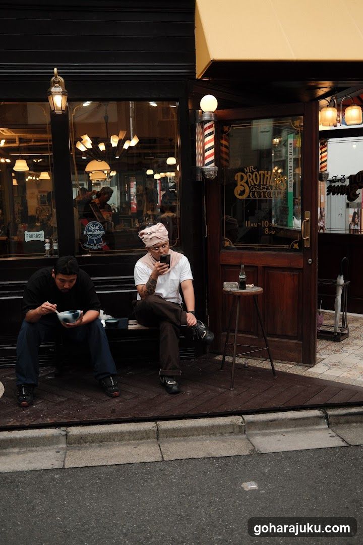 A barbershop storefront featuring a wooden entrance and glass windows displaying interior lighting and barber tools. Two individuals sit outside on the sidewalk; one eats from a bowl while the other uses a phone. A vintage-style barber pole sign is mounted near the door, along with illuminated lanterns above the window.