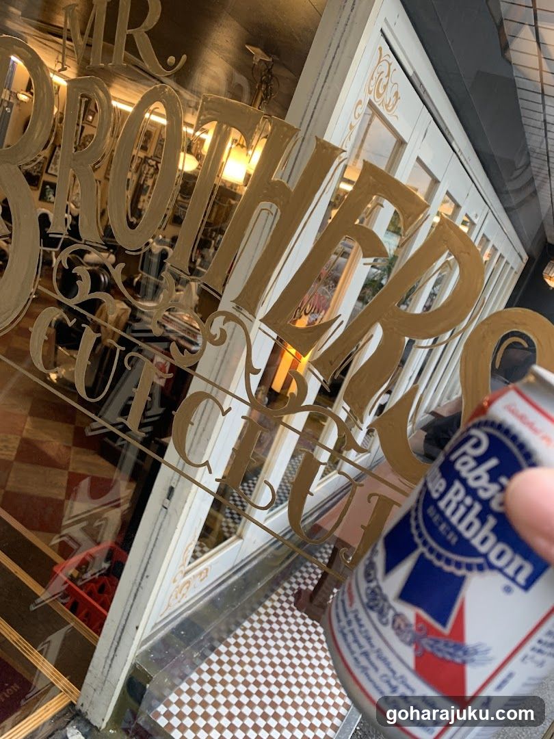 A close-up view of the storefront entrance featuring large gold lettering on glass reading 'MR BROTHERS CUT SHOP'. A hand holds a Pabst Blue Ribbon beer can in the foreground, partially obscuring the sign. The interior shows barber chairs and mirrors visible through the glass.