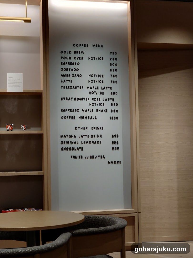 A vertical frosted glass menu board with black lettering displaying coffee and other drink options, including Gold Brew, Espresso, Latte, Maple Latte, Strawberry Rose Latte, Maple Shake, Coffee Highball, Matcha Latte Drink, Original Lemonade, Chocolate, Fruits Juice/Tea, and a S'more option.