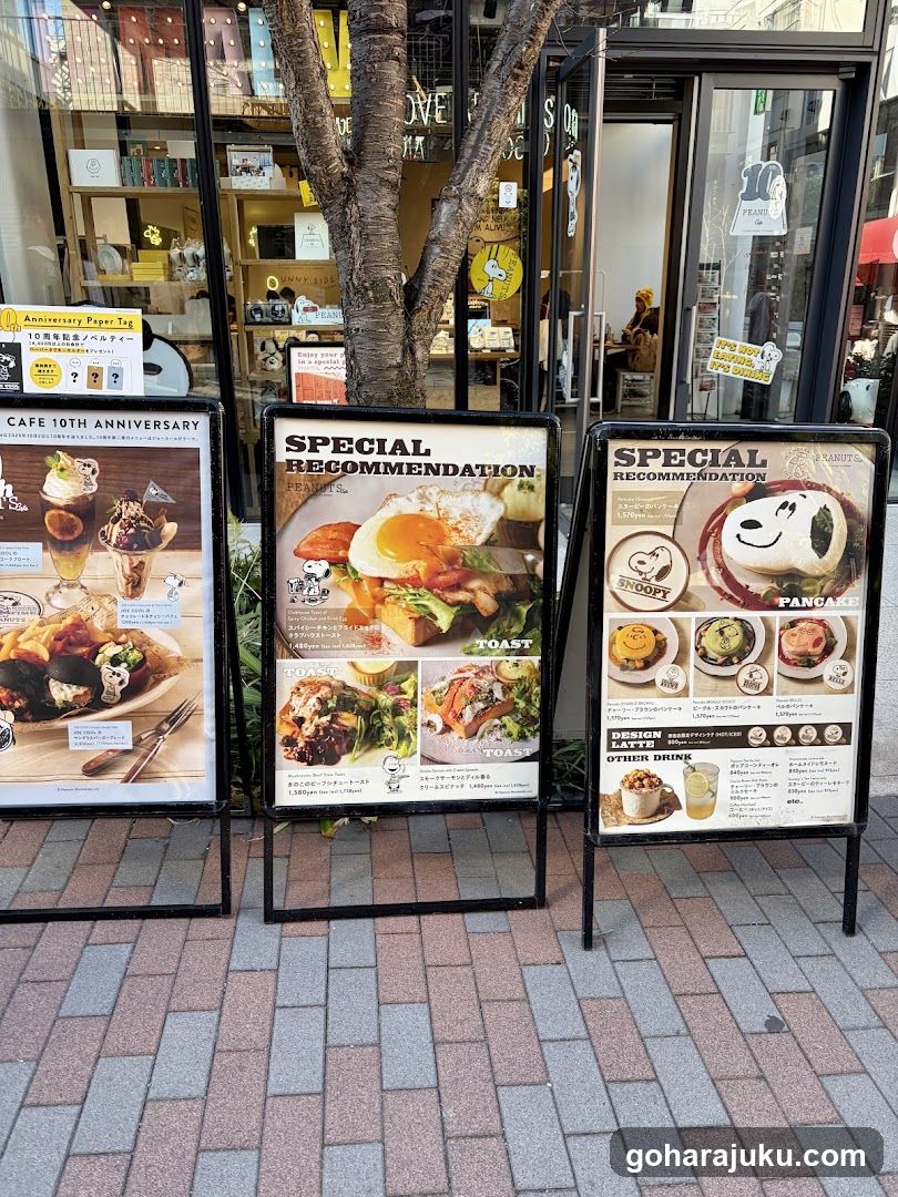 A cafe storefront featuring glass doors, a tree trunk in front, and three large menu boards on the sidewalk. The left board shows desserts and toast; middle highlights breakfast items like eggs and toast; right features character-themed pancakes and drinks. A small yellow sign mentions '10th Anniversary'. Interior shelves are visible through windows.