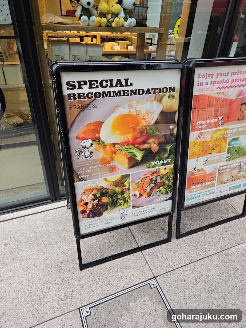 A photo of the exterior entrance showing two freestanding A-frame signs on a tiled sidewalk. The left sign advertises 'SPECIAL RECOMMENDATION' with images of food including toast and eggs, featuring Japanese text and English labels like 'TOAST'. The right sign promotes private rooms in colors (pink, yellow, green, white, orange) with room plans visible inside the glass window behind the signs.
