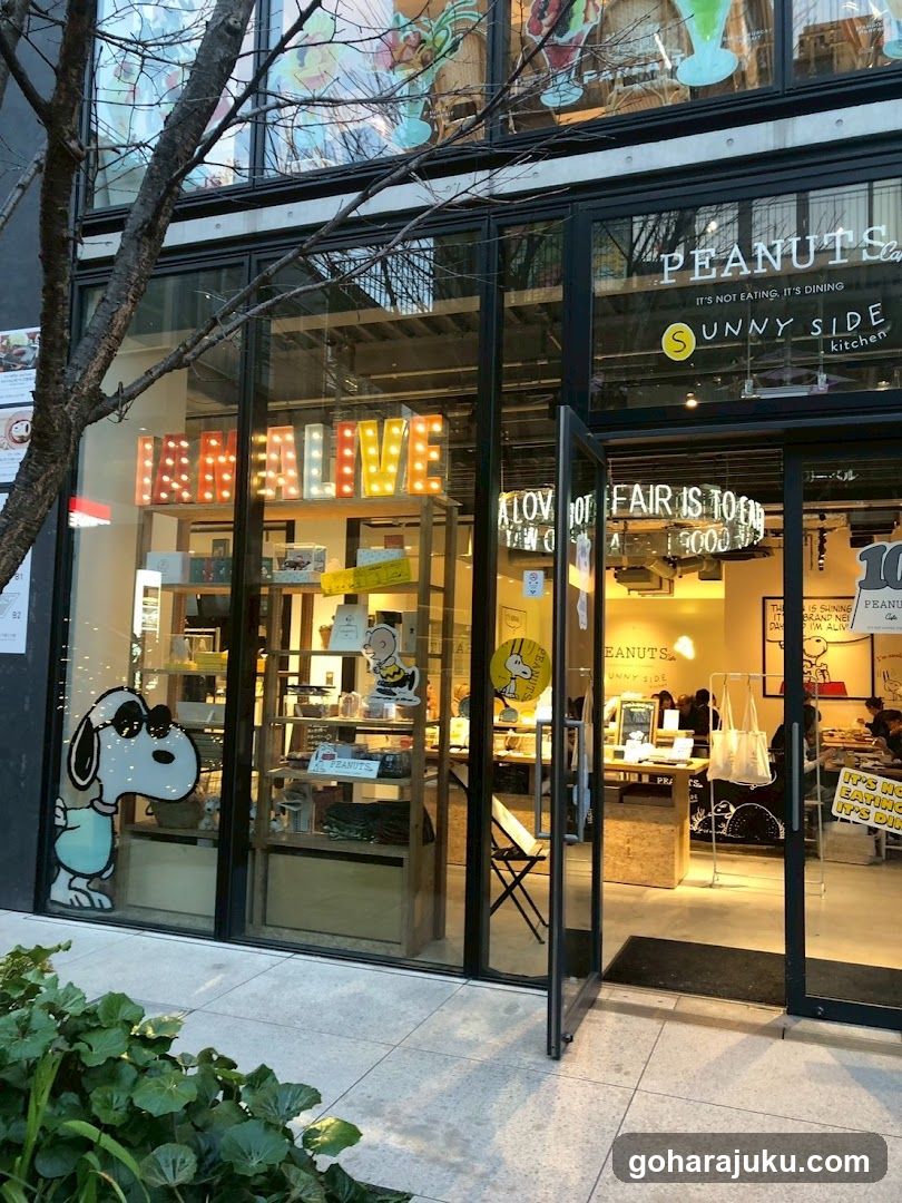 Storefront facade with large glass windows and doors, featuring illuminated 'PEANUTS' signage above the entrance. Snoopy character decal on left window. Interior displays shelves of merchandise, tables with seating, and hanging lights. Signage includes 'IT'S NOT EATING, IT'S DINING' and '100% FAIR IS TO BE'.