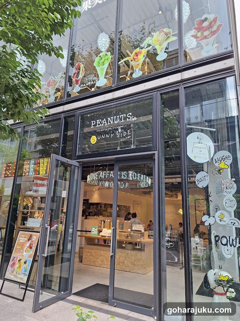 A modern glass-front cafe with visible signage reading 'PEANUTS Cafe' and 'SUNNY SIDE'. Decorative window decals feature fruit illustrations and Snoopy-themed elements including a snowman figure and speech bubbles. A standing menu board is placed outside near the open double doors, displaying colorful drink options inside.