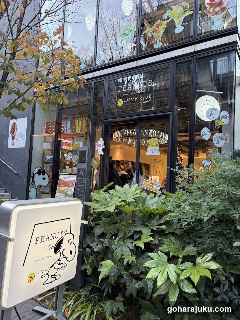 A modern glass-front cafe with black frames, featuring Snoopy and Woodstock decals on windows. A white signboard in front displays the 'PEANUTS' logo and a cartoon dog. Inside, customers are visible near the counter. Greenery surrounds the entrance.