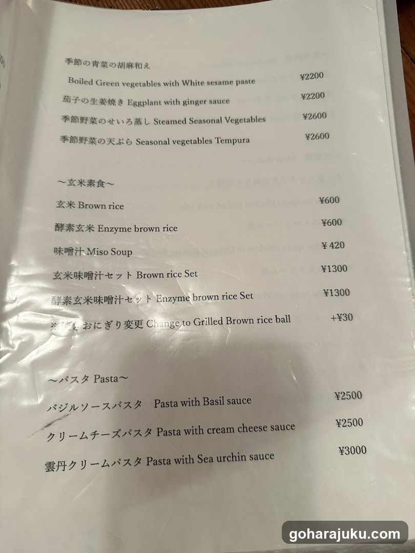 A printed menu page from a Japanese restaurant, featuring sections on seasonal vegetables, brown rice sets, miso soup, and pasta dishes with prices in yen.