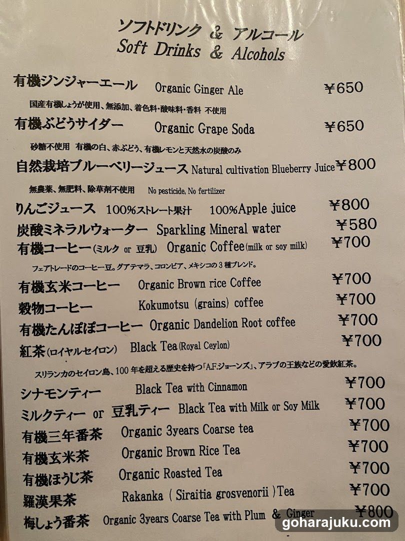 A printed menu listing soft drinks and alcoholic beverages with prices in yen, featuring items like Organic Ginger Ale, Organic Grape Soda, Natural Cultivation Blueberry Juice, Apple Juice, Sparkling Mineral Water, various coffee types (including organic brown rice and roasted tea), Black Tea with Cinnamon, Milk Tea or Soy Milk, Rakanka (Siraitia grosvenorii) Tea, and Organic 3years Coarse Tea with Plum & Ginger.