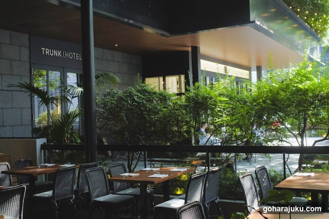Outdoor seating area with wooden tables and chairs, surrounded by lush greenery. A sign reading 'TRUNK (HOTEL)' is visible on the building facade.