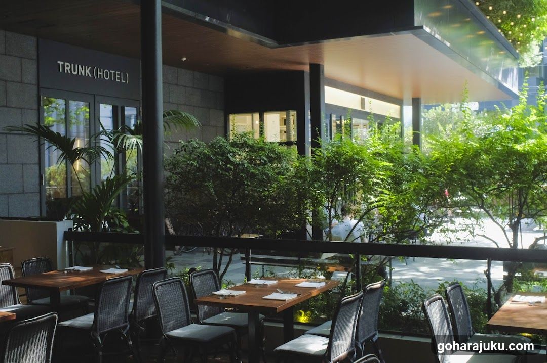 Photo of the physical exterior entrance/storefront facade with visible signage 'TRUNK (HOTEL)' on a modern building featuring large windows and greenery surrounding outdoor seating areas.