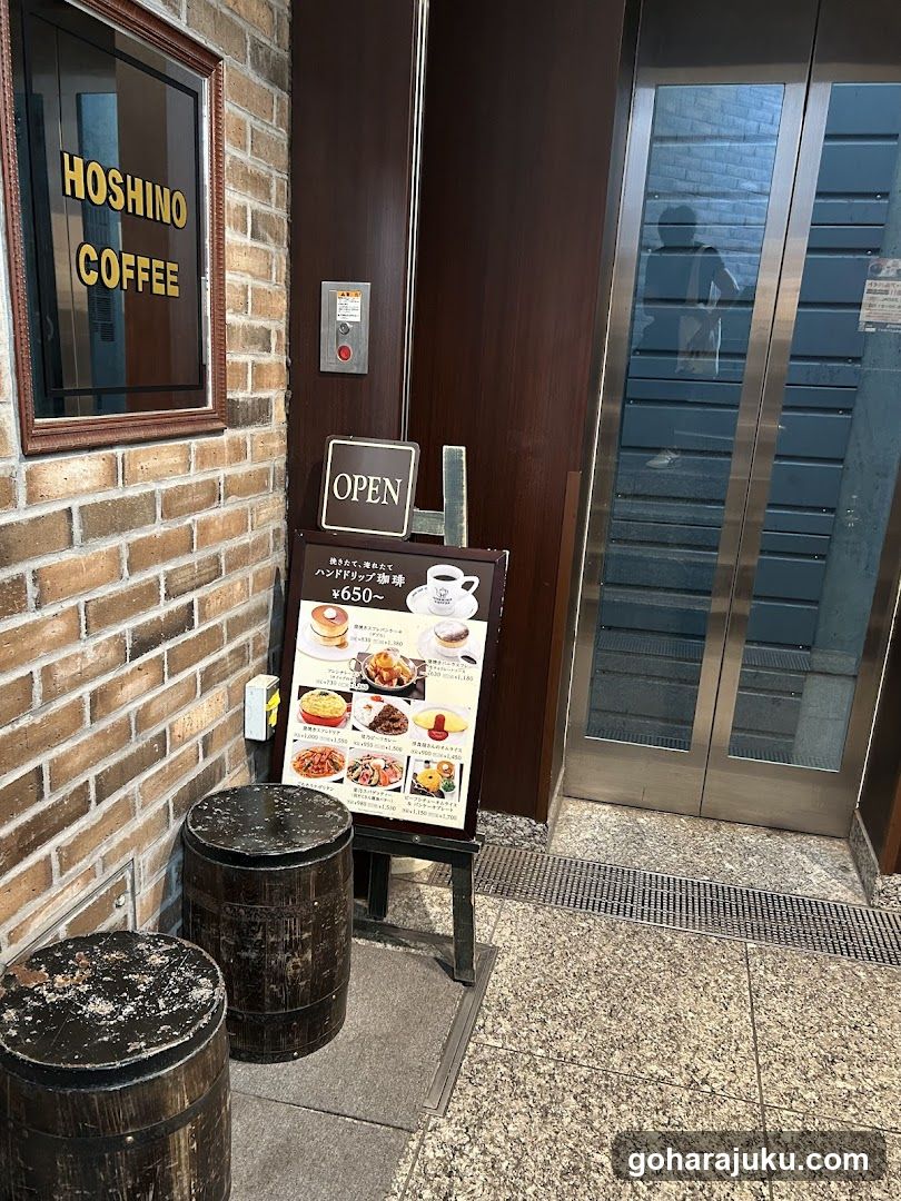 Exterior view of Hoshino Coffee storefront featuring brick wall with framed signage, wooden door, glass elevator doors, and a menu board on stand near entrance.