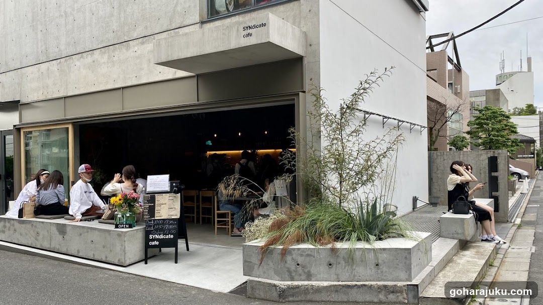 A modern concrete storefront with large glass windows and a sign reading 'SYndicate cafe' above the entrance. People are seated on benches outside, enjoying the space. A menu board stands near the door.