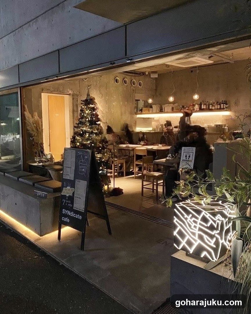 A cozy cafe exterior featuring a lit Christmas tree inside, warm lighting, and patrons dining at tables. A signboard outside displays menu information and the word 'OPEN'. The interior has exposed walls and shelves stocked with bottles.