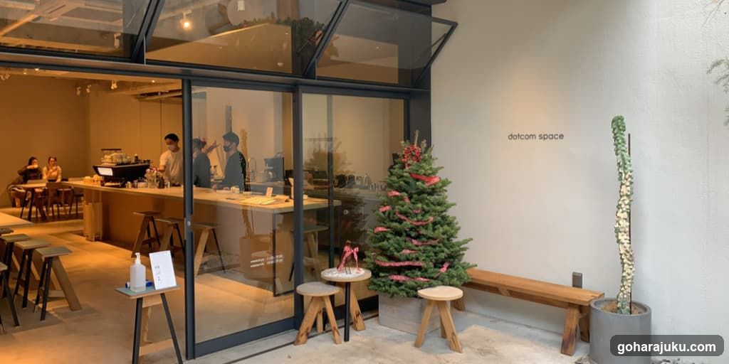 A modern coffee shop with glass facade, visible interior staff and customers, a small decorated Christmas tree outside, wooden stools, and a sign reading 'dolcom space' on the wall.