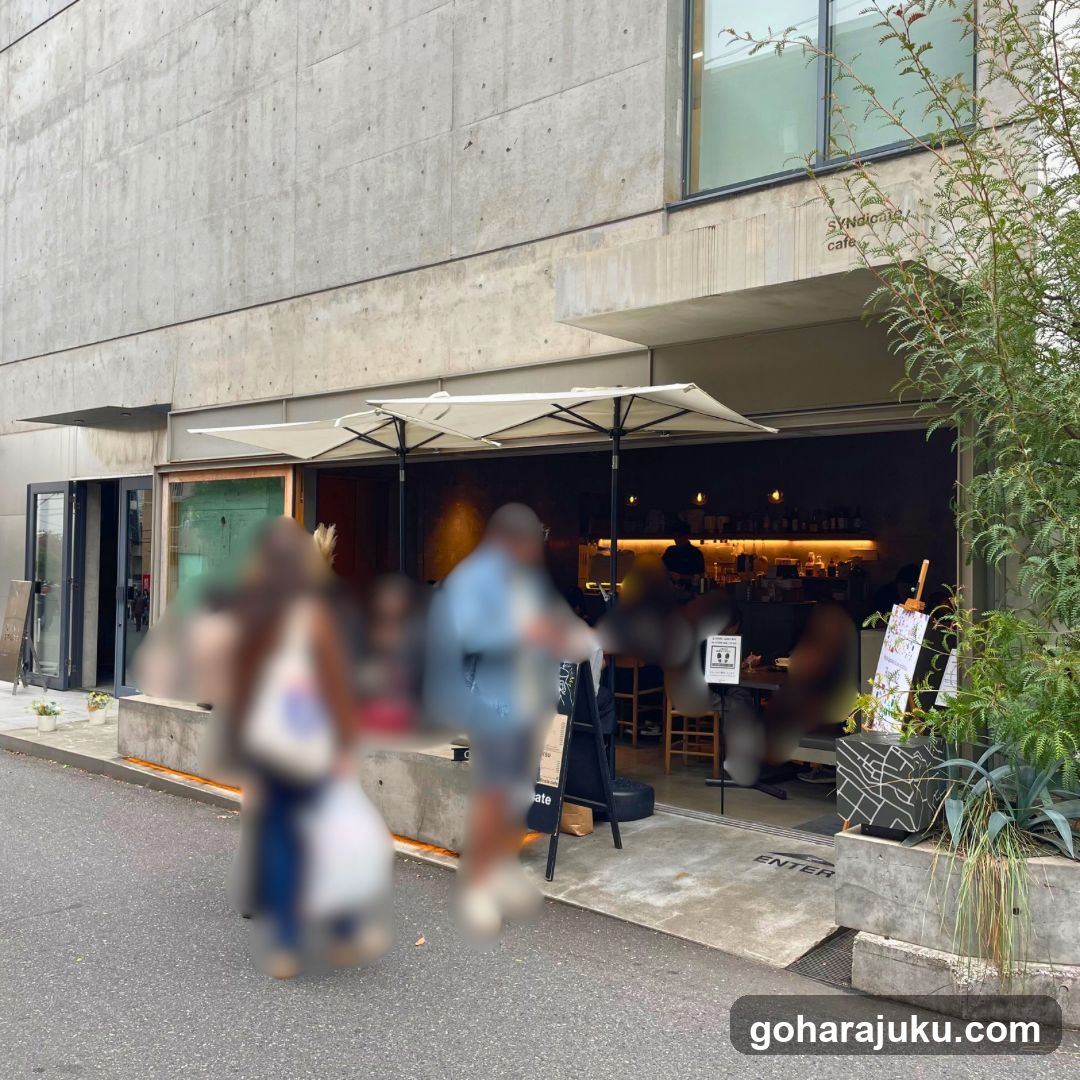 Photo of the physical storefront facade showing a concrete building with large glass windows and doors. A white umbrella shades the entrance area where people are standing. Signage reading 'Syndicate cafe' is visible on the upper right wall. No menu boards or price lists are present in this exterior shot.
