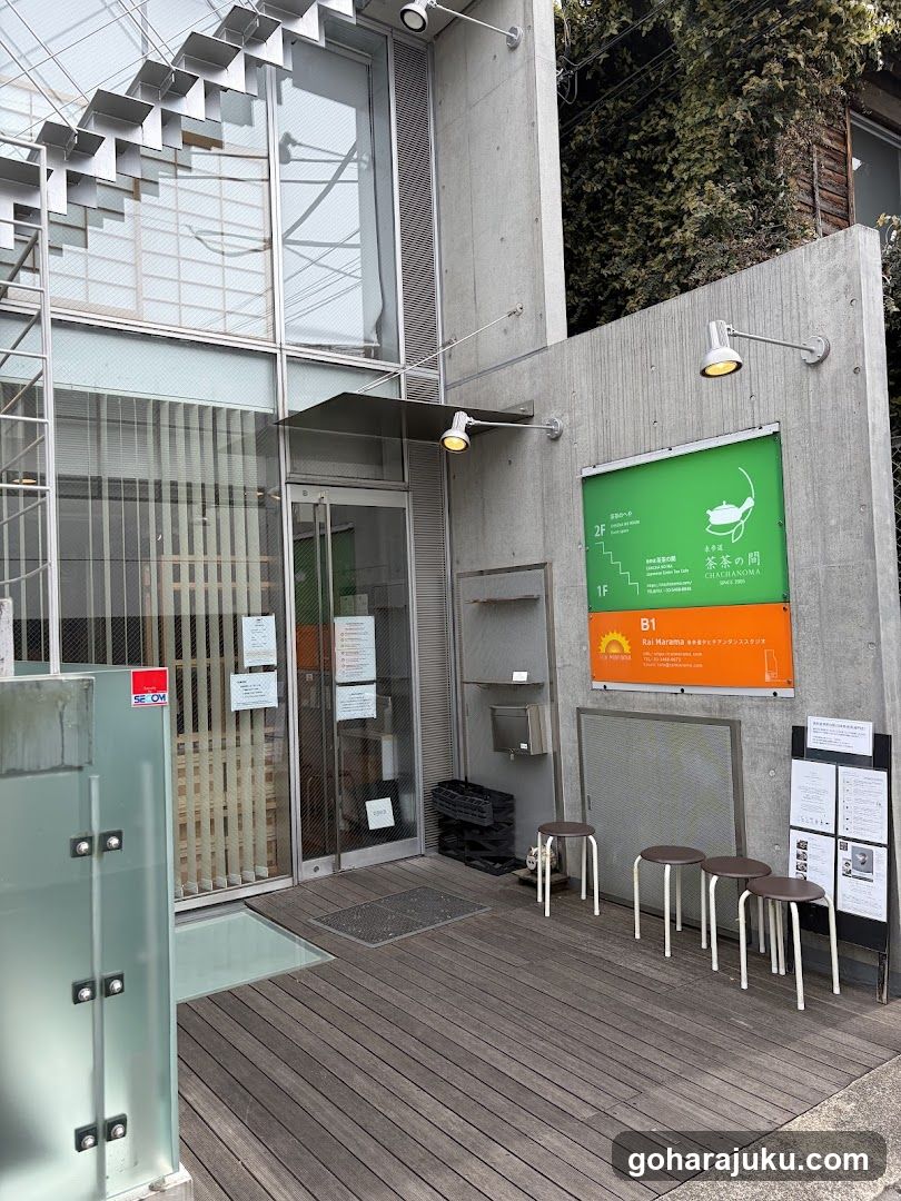 The image shows the exterior entrance of a building with glass doors and a wooden deck area. There is a green sign on the wall indicating '2F' and '1F', along with an orange section labeled 'B1'. Several stools are placed near the entrance, and there are notices posted on the walls.