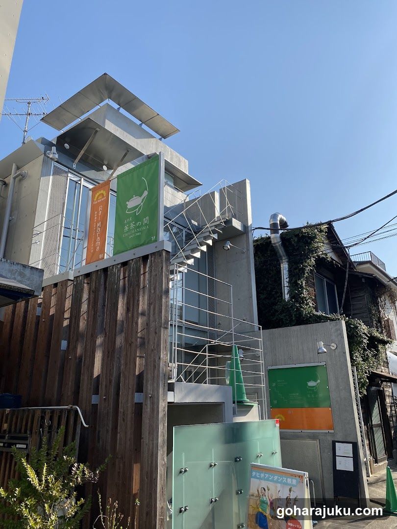 A modern building exterior featuring hanging banners, one green and one orange, attached to the facade. The structure includes a wooden fence in front, metal railings on an external staircase, and glass panels. A poster board is visible near the entrance.