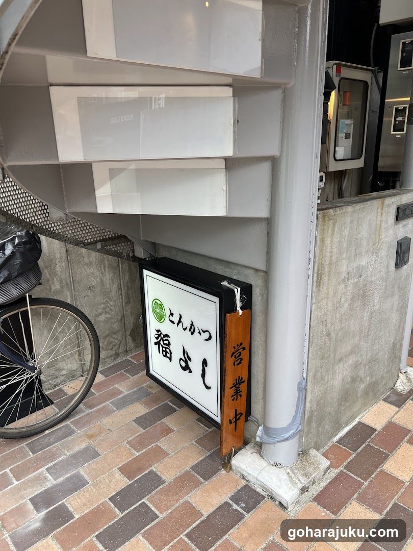 A shop front featuring a sign with Japanese characters 'Fukuyoshi' and 'Don Katsu', indicating it is open for business. A bicycle wheel is visible to the left, and the building has a modern design with concrete walls and metal railings.