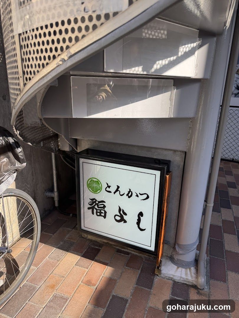 A signboard with the text 'とんかつ' (Tonkatsu) and '福よし' (Fuyoshi), featuring a lime graphic, is mounted on the wall beneath a spiral staircase.