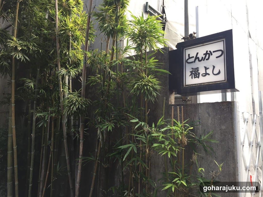 A narrow alleyway entrance featuring tall bamboo plants lining the left side, leading to a building facade. A black-framed signboard mounted on the wall displays Japanese characters 'とんかつ' (tonkatsu) and '福よし' (fuyoshi), indicating a tonkatsu restaurant. The background includes concrete walls and part of a white lattice gate.