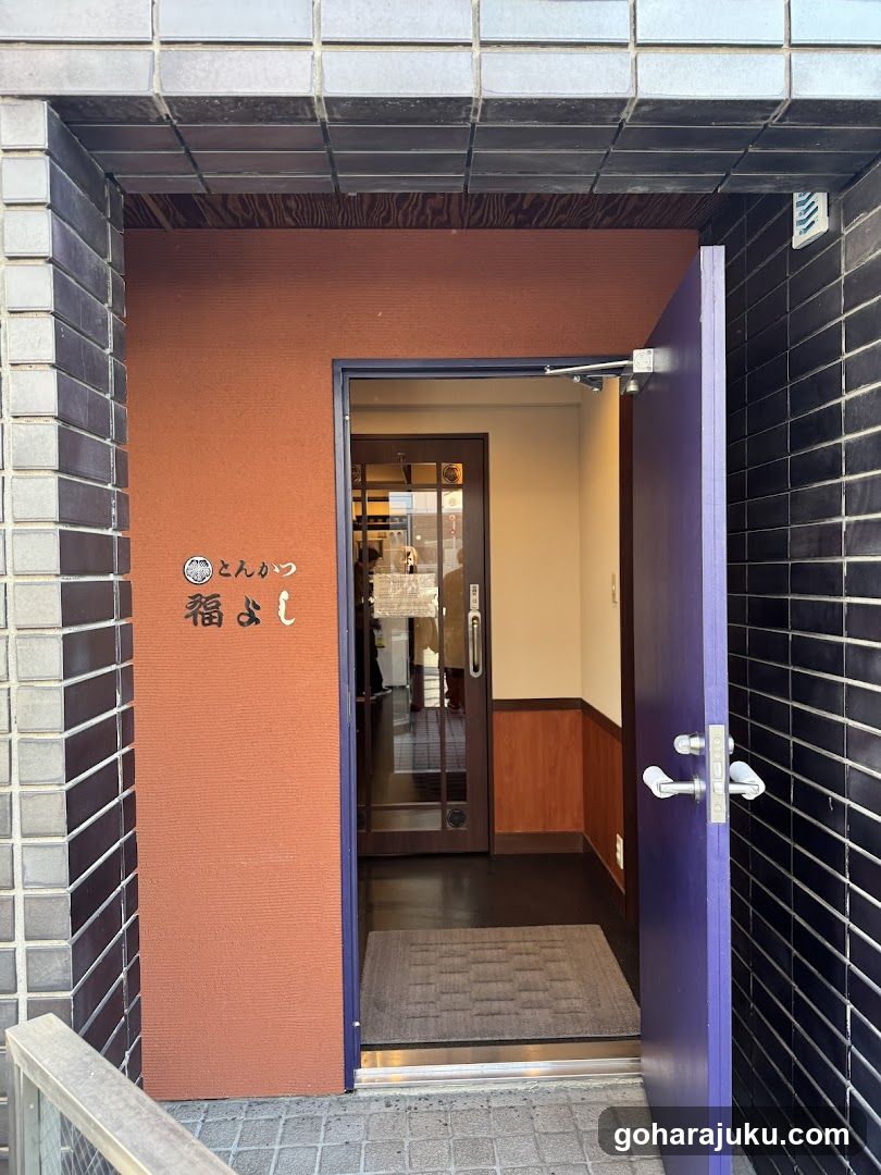 The image shows the exterior entrance of a building with an orange wall and black tiled sides. A purple door is open, revealing a glimpse into the interior. On the orange wall, there are signs in Japanese characters and symbols, indicating it might be a shop or restaurant.