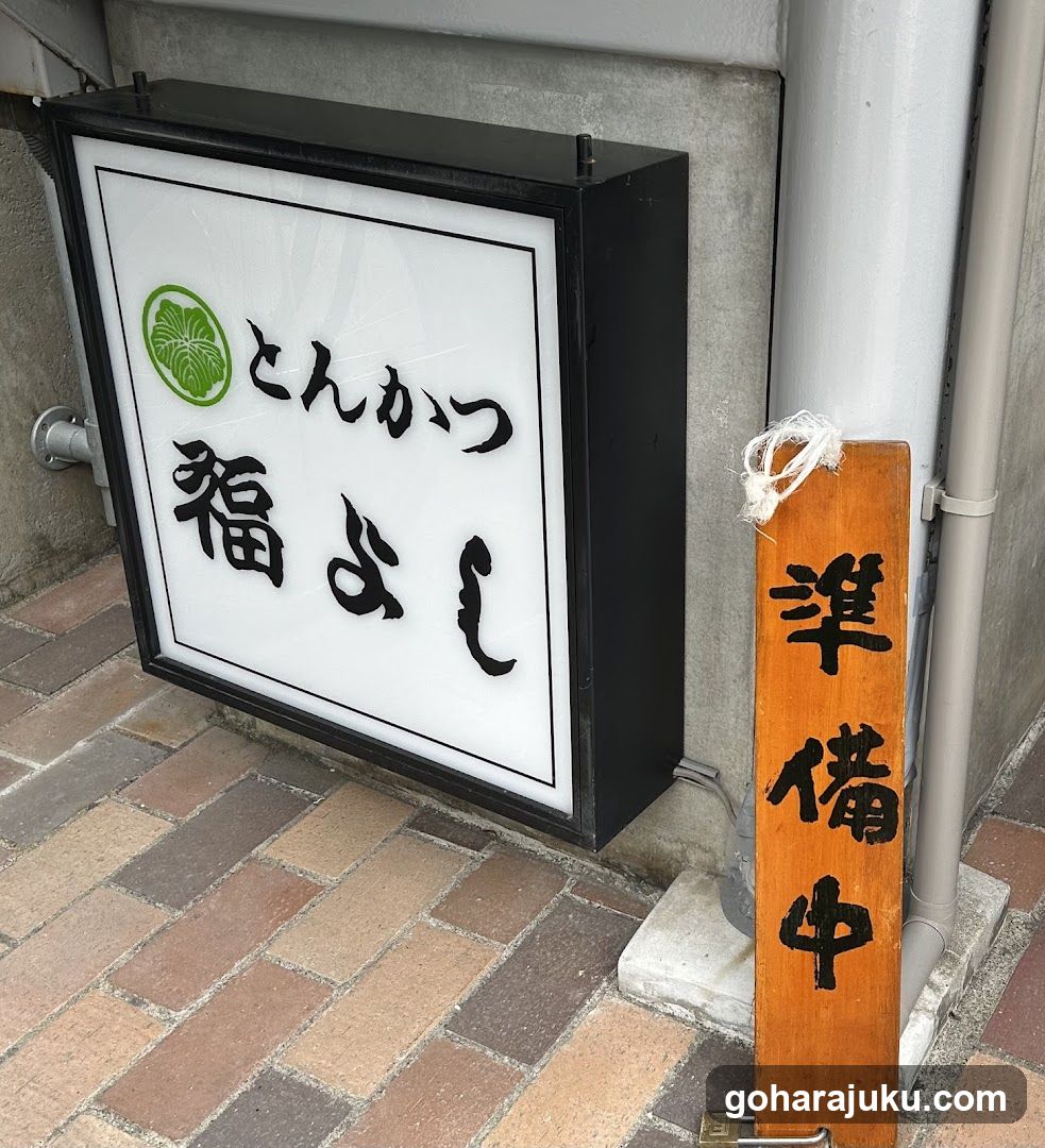 A lit rectangular sign with Japanese characters 'とんかつ' (tonkatsu) and '福よし' (Fuyoshi), alongside a green circular logo resembling a leaf or flower pattern. Next to it is an orange wooden hanging sign reading '準備中' (preparation in progress). The signs are mounted on the exterior wall of a building, above a brick-paved sidewalk.