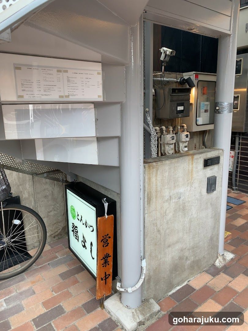 The image shows the exterior of a shop with a menu board mounted on the wall, displaying various items and prices. A bicycle is parked nearby, and there are utility meters visible in the background.