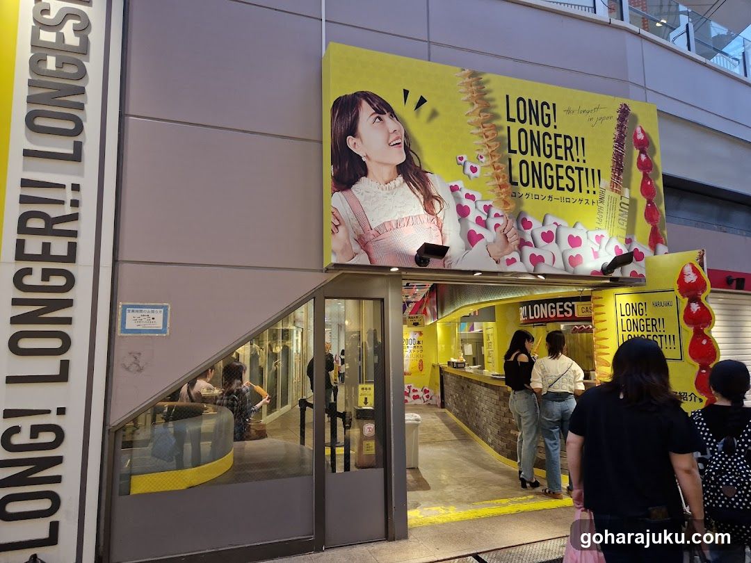 A shop entrance with a large yellow billboard featuring a smiling woman and heart-shaped products. The sign reads 'LONG! LONGER!! LONGEST!!!'. People are standing near the counter, which has similar branding. A vertical sign on the left also displays the same slogan.