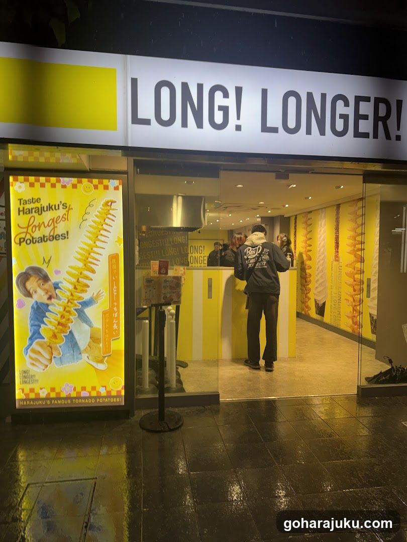 A shop front with a large sign reading 'LONG! LONGER!' above the entrance. A poster on the left advertises Harajuku's longest potatoes, showing a child holding a spiral potato. Inside, customers are visible near the counter, and yellow-themed walls feature images of long spiral potatoes.