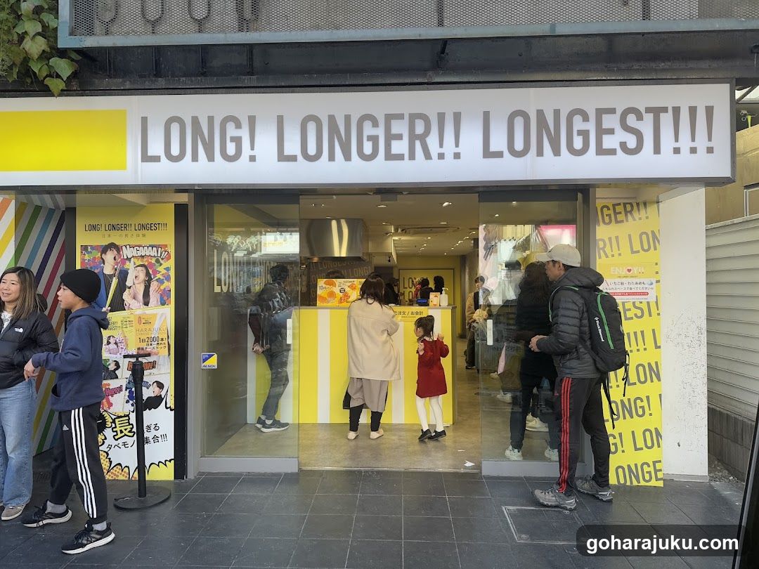 A storefront with a large sign reading 'LONG! LONGER!! LONGEST!!!'. The entrance features glass doors and windows, with yellow and white striped decorations inside. People are standing outside, some looking at posters on the wall.