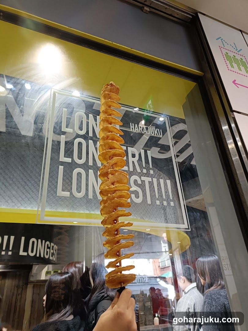 A hand holds a long spiral potato snack in front of a shop window displaying 'HARAJUKU' and repeated words like 'LONGER!!'. The storefront features yellow accents and visible reflections on the glass.