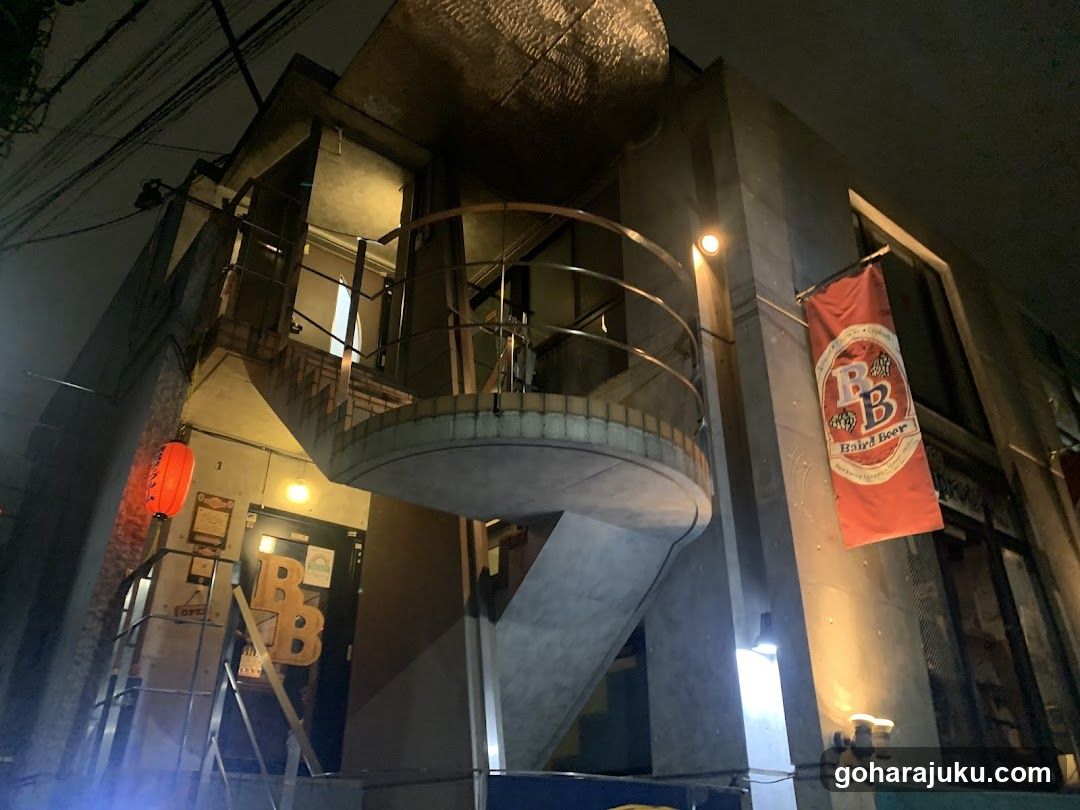 A nighttime photo of a building featuring an external concrete spiral staircase leading to an upper level. A red banner hangs on the right side displaying 'PB' logo and text below it. On the left, there is a doorway with large letters spelling out part of a name or brand. The scene includes ambient lighting from wall-mounted fixtures and a lantern hanging near the entrance.