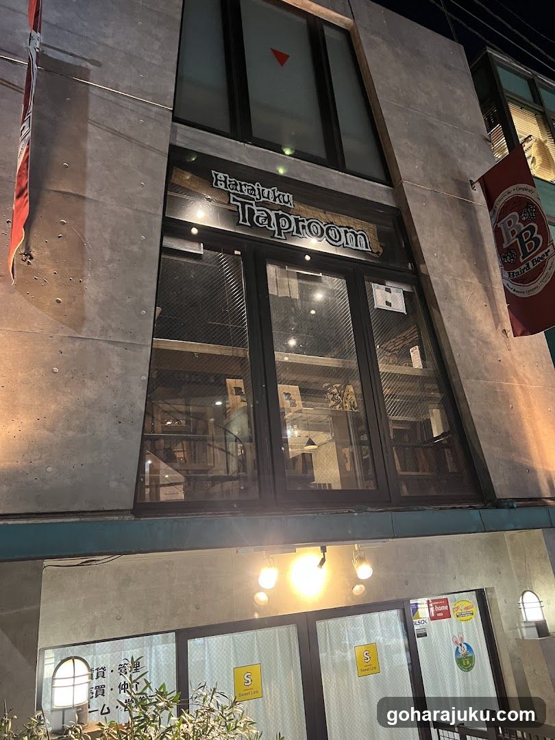 Night exterior shot of a concrete building facade with large glass windows and doors. Signage reads 'Hakainu Taproom' above the entrance. A red flag hangs on the right side. Interior lights are visible through the glass, showing tables and chairs.