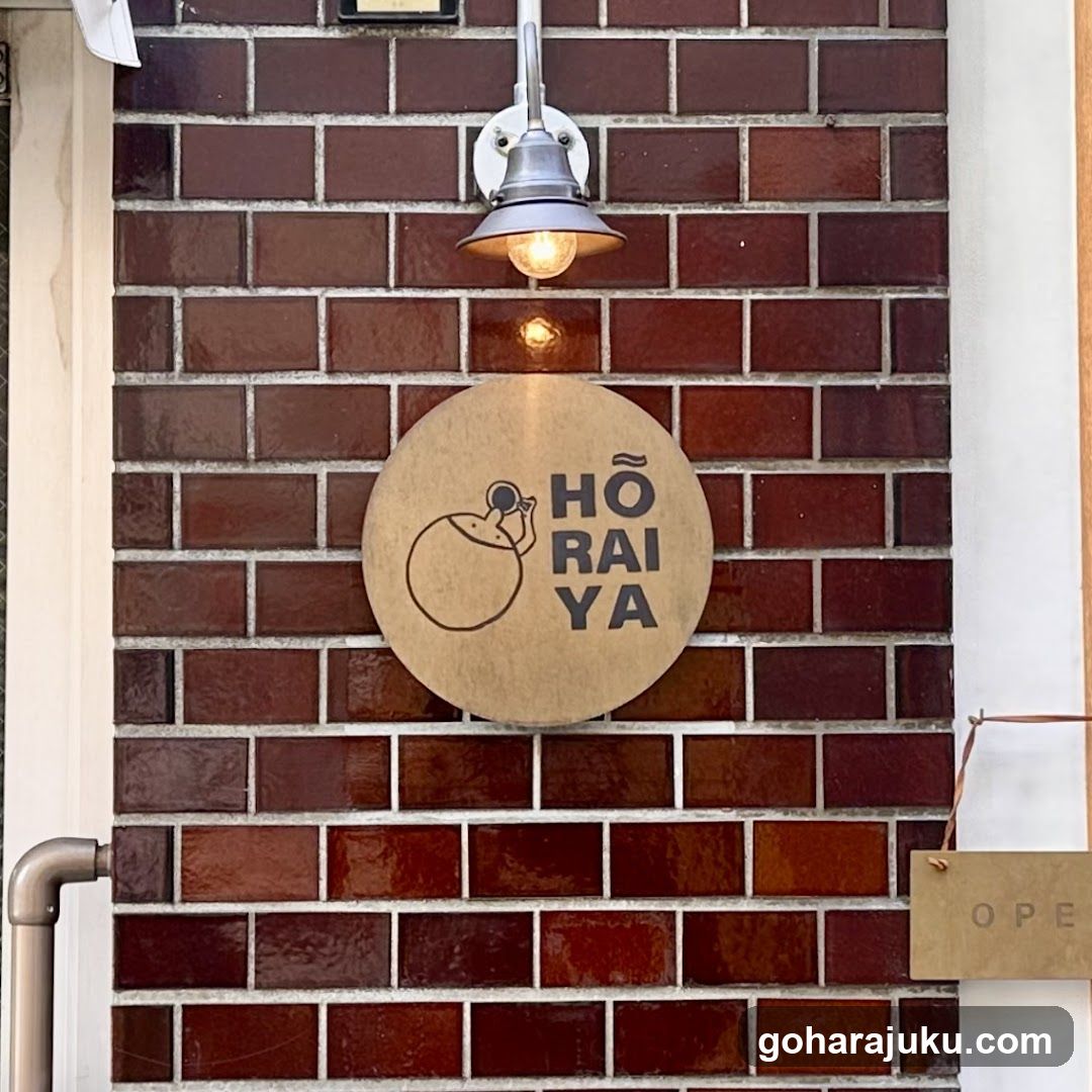 A circular wooden sign with the text 'HŌ RAI YA' and a simple line drawing of a person holding a camera is mounted on a red brick wall. Above the sign, an industrial-style light fixture illuminates the area. To the right, part of another sign reading 'OPEN' is visible.